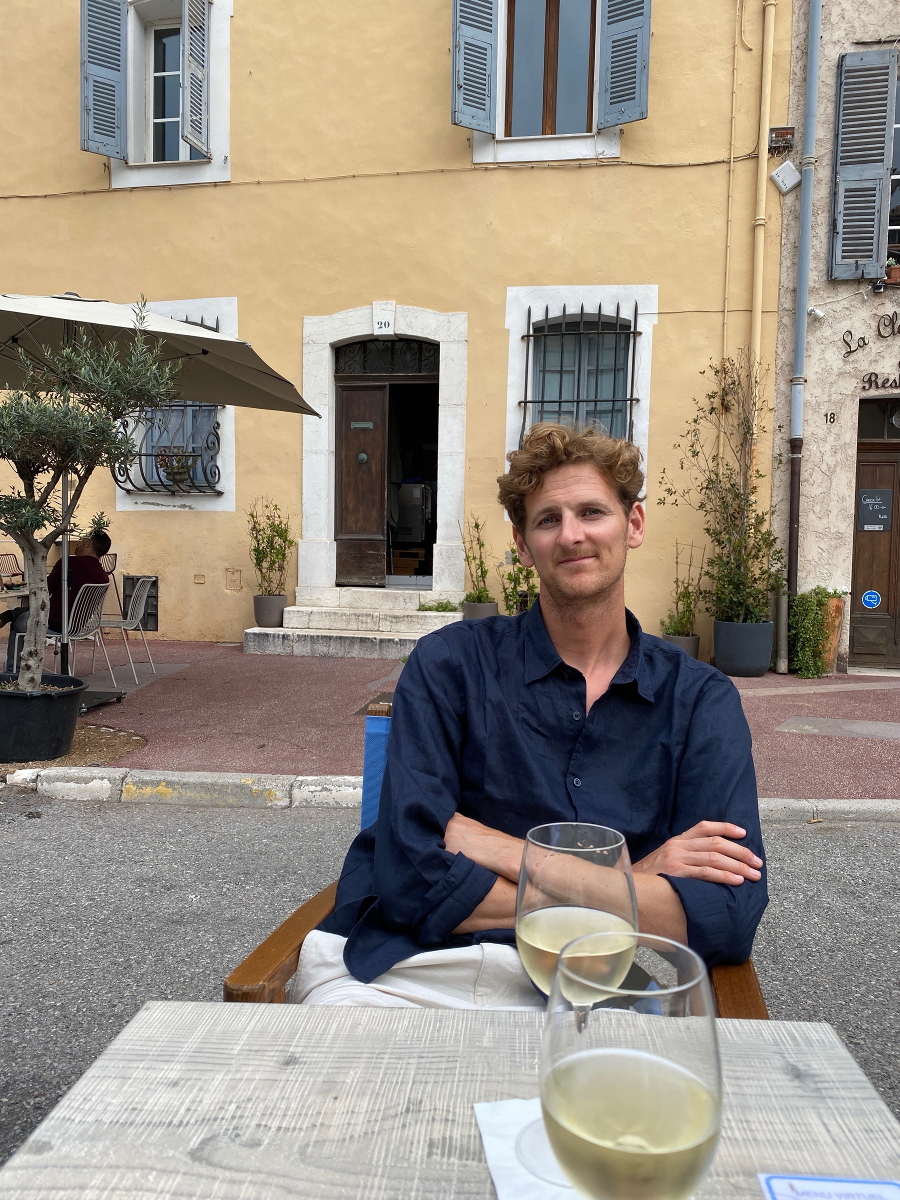 Ed at a café table on a sunny village square, arms folded, a glass of white wine on the table.