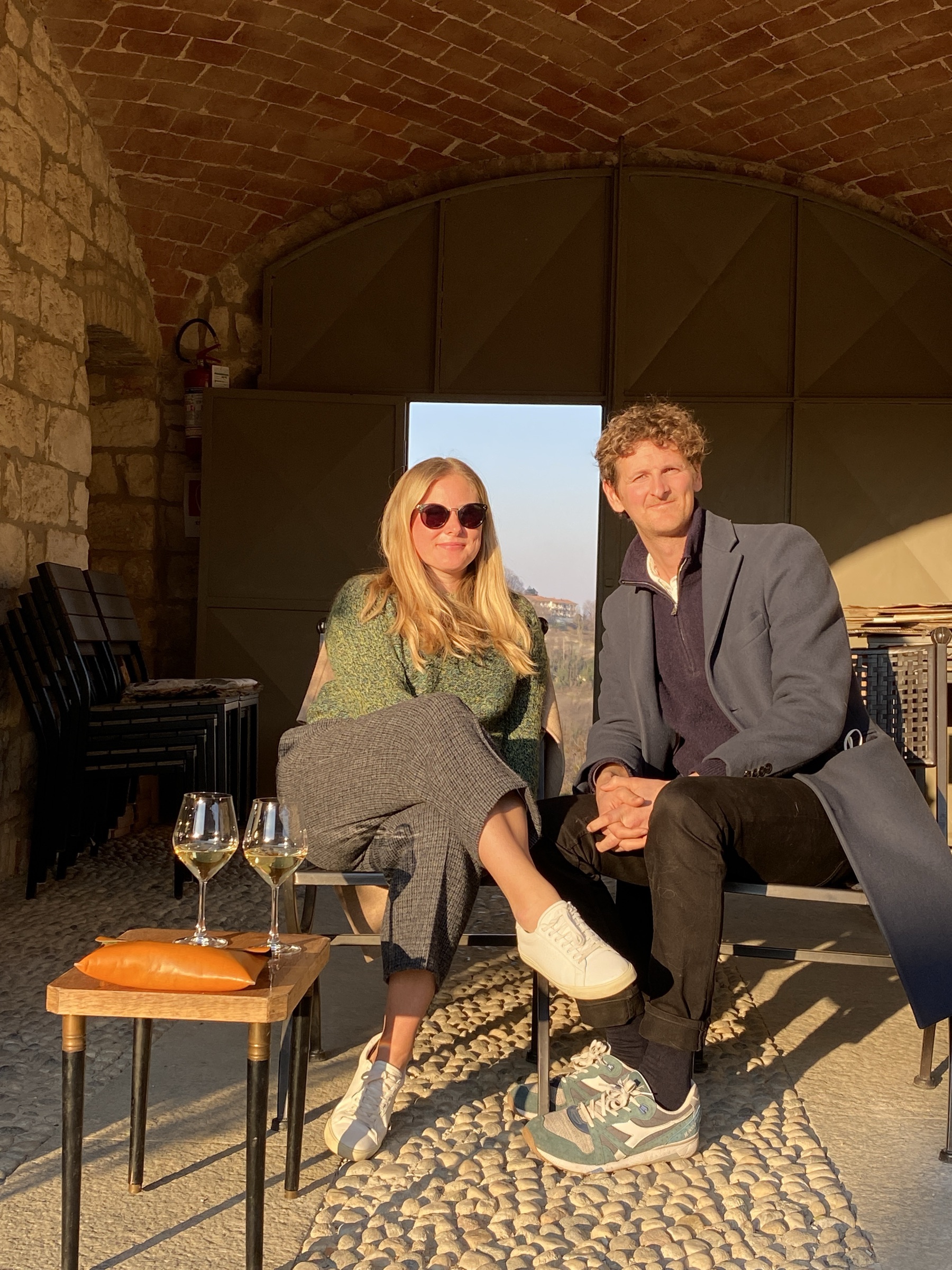 Ed and Emelie sitting side by side in late sun under a brick vault, two glasses of wine on a small table.