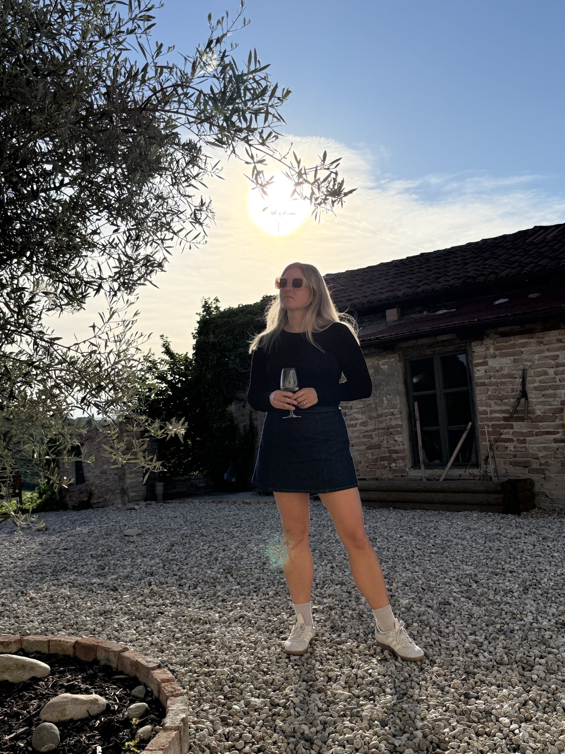 Emelie standing on a gravel courtyard in evening sun in front of an old brick house, glass of wine in hand.