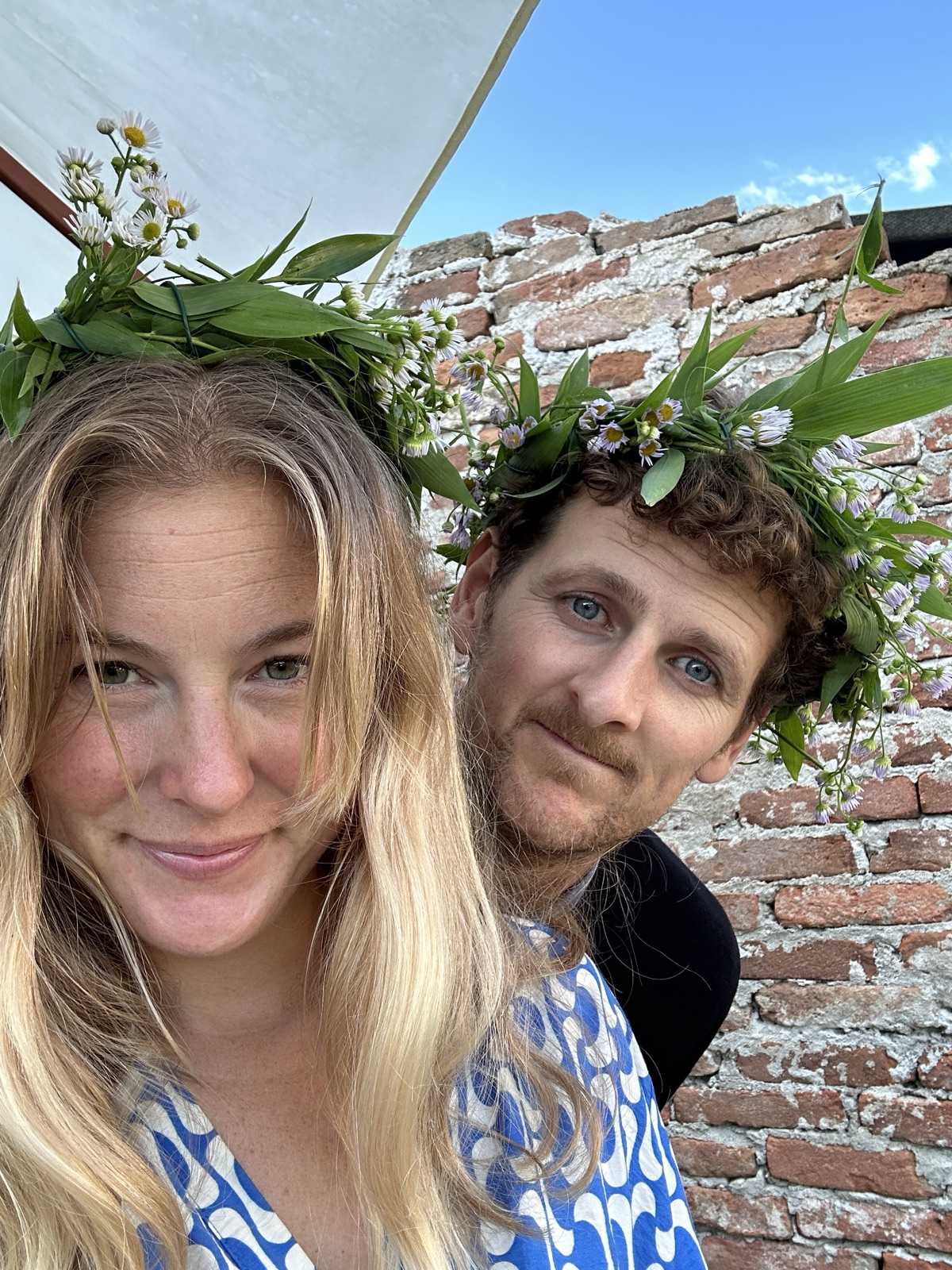 Emelie and Ed in flower crowns, smiling in front of an old brick wall.